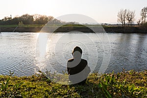 A hooded man sitting by a river, back to camera, with a feeling of calm, solitude and thinking