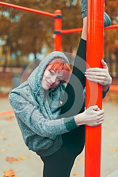 Hooded parkour woman warming up at playground pillars and doing splits in park at autumn