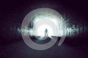 A hooded silhouetted figure standing on the edge of a tunnel looking out onto a bright overexposed light photo
