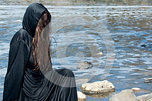 Hooded Woman with dark long hair in black robes in front of the Lake. Witches. Halloween and Gothic. Witchcraft and magic.