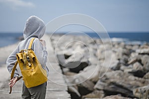 A hooded woman puts on a backpack while standing on an ocean pier. Back view.