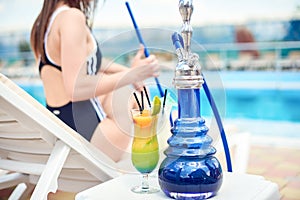 Hookah or shisha or kalian and a tropical summer refreshing alcoholic coctail in front of swimming pool and a beautiful