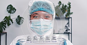 Hope fighting coronavirus. Serious multiethnic female lab scientist in protection suit showing tray with vaccine flasks.