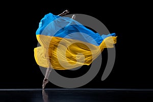 Hope. Young graceful classic ballerina dancing with cloth painted in blue and yellow colors of Ukraine flag on dark