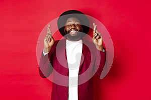 Hopeful african american man making wish, holding fingers crossed for good luck and smiling optimistic, standing against