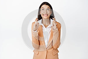 Hopeful businesswoman waiting with excitement, cross fingers and wishing, looking up in anticipation, standing in suit