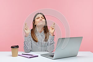 Hopeful dreamy woman office worker sitting at workplace in anticipation of business event, crossing her fingers and wishing for