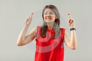 Hopeful and nervous. Portrait of worry brunette young woman in red shirt standing, clenching teeth, crossed fingers and hope to be