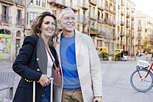 Hopeful senior man and woman standing with a suitcase happy and relaxed in old town. Mature couple hugging together with