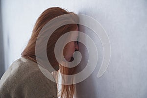 Hopeless and depressed young redhead woman leans her forehead on wall at home