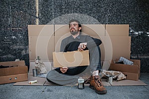 Hopeless and homeless person is sitting on cardboard on the ground and holding a piece of cardboard. He looks tired and