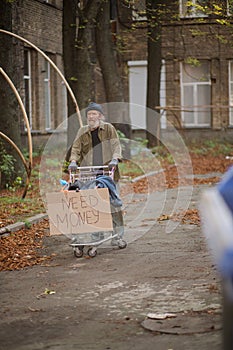 Hopeless old tramp with cart and board with sign need money.