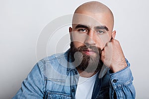 A horizontal close-up of handsome bald man having charming dark eyes, thick black eyebrows and beard wearing jean shirt holding hi