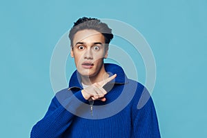 horizontal close-up portrait of a surprised man standing on a light blue background in a blue zip-up jacket and pointing