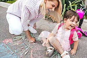 Horizontal image of happy little girl and mother laughing and drawing with chalks on the sidewalk. Caucasian female play together