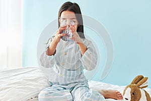 Horizontal image of a little girl drinking fresh water in the bed at home. Cute preschool kid holding glass of pure water in the