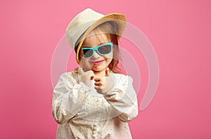 Horizontal image of little girl in straw hat and sunglasses, shows a thumb up, glances at you, stands on isolated pink.