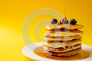 Horizontal image of maple syrup pouring onto pancakes and blueberries, on yellow with copy space