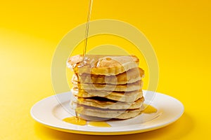 Horizontal image of maple syrup pouring onto stack of pancakes, yellow background and copy space