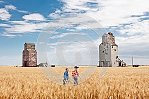 Two cowboy farmers walking in wheat field by elevators.