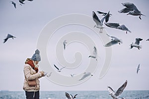 Horizontal image of young beautiful woman feeds seagulls on the sea. Pretty female wearing coat, scarf, hat watching flying