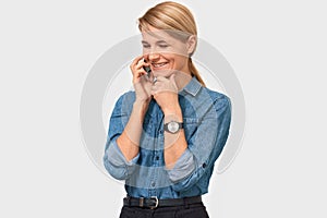 Horizontal indoor image of young pretty blonde woman in denim shirt smiling and talking on smart phone to her friend