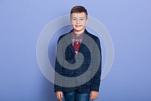 Horizontal indoor shot of positive sweet child standing isolated over lilac background, wearing jeans, plaid shirt and dark jacket