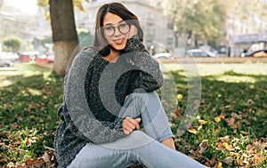 Horizontal outdoor image of pretty smiling student female looking to the camera, sitting on green grass in the city park, wearing