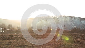 Horizontal panorama, burning grass near forest with lots of smoke next to wooden houses at sunset against mountain