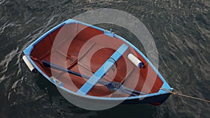 Small wooden fishing boat with a single blue oar inside, tied up near a pier against dark, murky ocean water