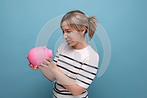 horizontal photo of an attractive young woman in a casual outfit with a piggy bank of coins on a blue background with