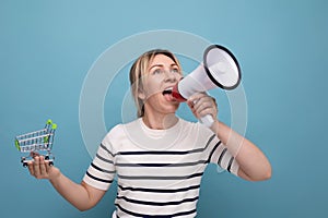 horizontal photo of blonde attractive young woman with megaphone screaming news holding grocery cart on blue background