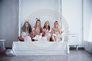 Horizontal photo. Front view. Sitting on the luxury white bed with balloons and bunny ears. Conception of bachelorette