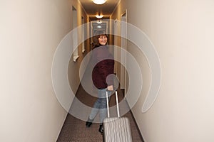 Horizontal photo of a woman, a tourist, walking along a long corridor of the hotel with a suitcase past the door, tourist concept
