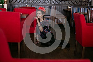 Horizontal photo of a young beautiful woman with bright makeup and flowers in her hair who is sitting on the floor sadly looking