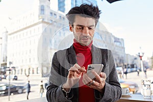 Horizontal photo of young handsome dark haired man dressed in formal clothes typing message on his mobile phone while standing