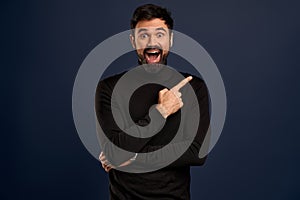 Horizontal portrait of bearded cheerful man has smile, wears trendy sweater, indicates with fore finger at copy space for your