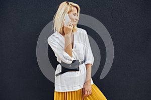 Horizontal portrait of blonde business woman smiling and standing against gray building`s wall while talking on cellphone.