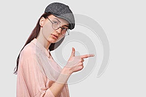 Horizontal portrait of candid young woman wearing pink shirt and round transparent glasses, indicates with forefinger demonstrates