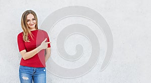 Horizontal portrait of cute woman wearing red sweater and jeans standing near white concrete wall poiting with her index finger at