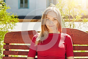 Horizontal portrait of fair-haired pretty young girl with freckles and blue eyes wearing red sweater sitting at bench in park admi