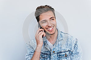 Horizontal portrait of fashionable male in jeans jacket has communication with friend via mobile phone. Handsome man speaking vie