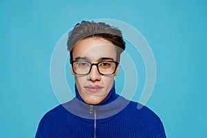 horizontal portrait of a handsome man in a blue zip-up sweater and black eyeglasses, standing on a light blue background