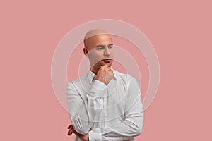 Horizontal portrait of joyful bald guy in white shirt with thoughtful expression holds hand above chin, looks seriously