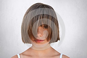 Horizontal portrait of little freckled female child covering her face with hair while poising against white concrete wall. Adorabl