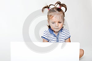 Horizontal portrait of little pretty girl focused into laptop computer, watches interesting cartoon or play games, has two pony ta