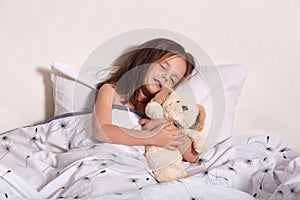 Horizontal portrait of little sweet cute child lying in her bed, closing eyes, sleeping soundly, having dreams, hugging her teddy