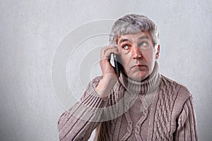 A horizontal portrait of mature man with gray hair and wrinkles dressed in warm sweater holding mobile phone on his ear looking as