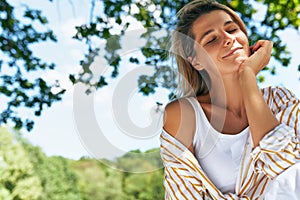 Horizontal portrait of a pretty young blonde woman smiling with eclosed eyes, enjoying the sunny day in the park. Beautiful female