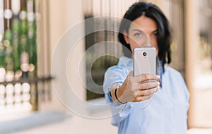 Horizontal portrait of pretty young woman makes selfie on smart phone, poses at camera while waiting her freinds at the park,